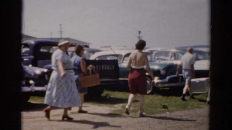 Adults carrying baskets walk near a line of parked 1950s cars, captured as casual 8mm home-movie footage in Catskill, New York.