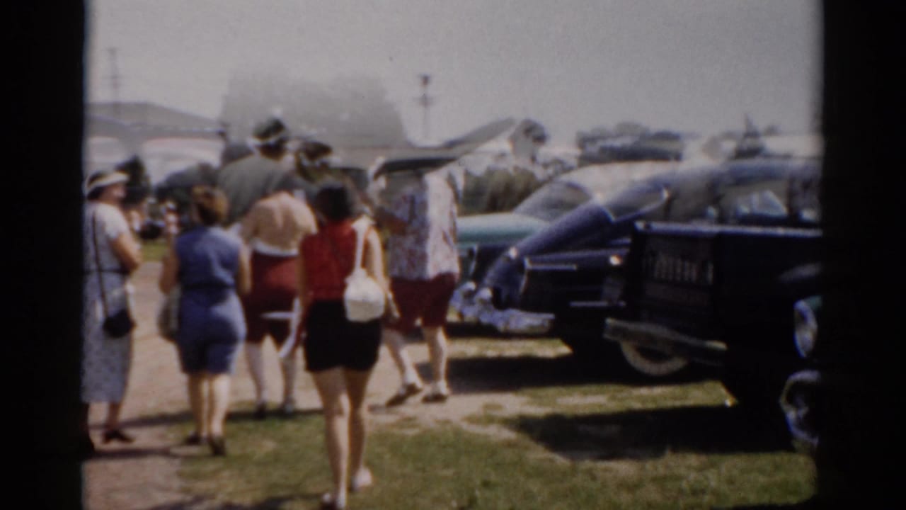 Group of adults walking away from camera past parked 1950s American cars on a sunny day.