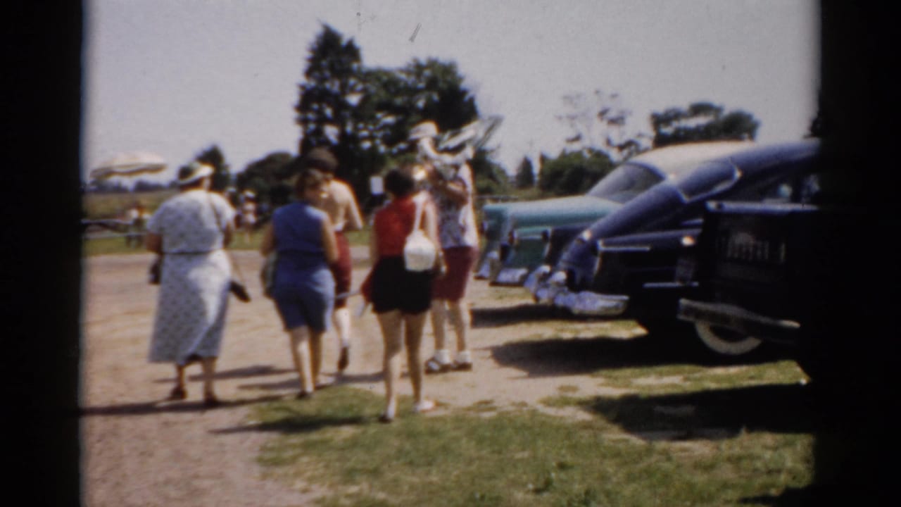 Women in summer skirts and shorts walking away from camera past a line of large chrome-detailed American cars, consistent with 1957 Catskill, New York home-movie footage.