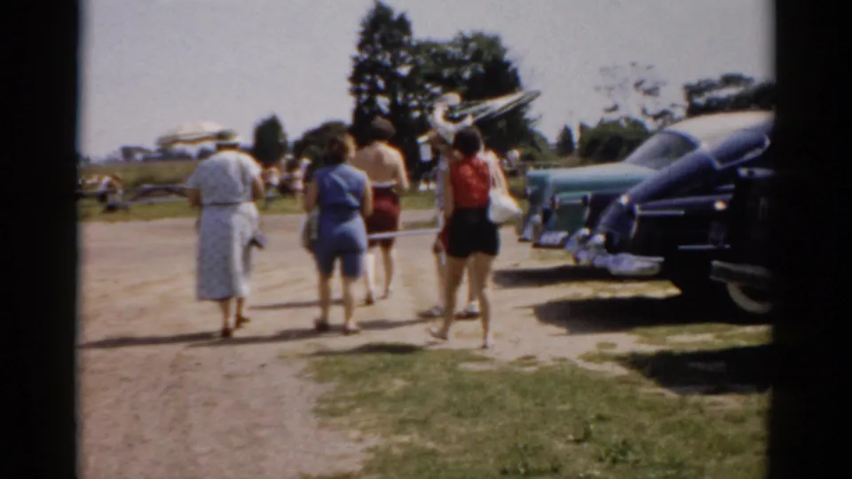 Group of adults, women prominent in skirts and shorts, walking away from camera in 1950s summer clothing beside a row of late-1950s American automobiles, Catskill, New York area.