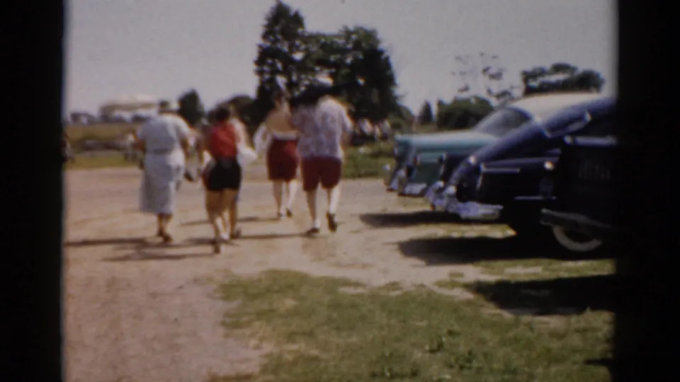 Continuing shot of adults in 1950s summer dress walking away from camera past a row of large American cars, trees and open sky visible behind, circa 1957, Catskill, New York.