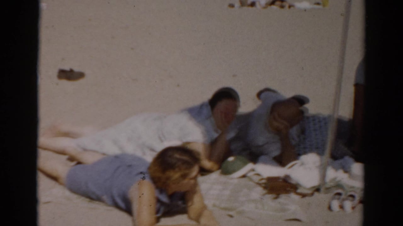 Woman with dark hair lying prone on a beach blanket, man reclining behind her, umbrella pole visible at center, sandy beach.