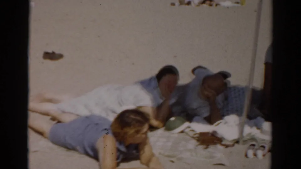 Woman lying on stomach on a plaid beach blanket, man reclining behind her; 8mm home movie, Catskill, New York, circa 1957.