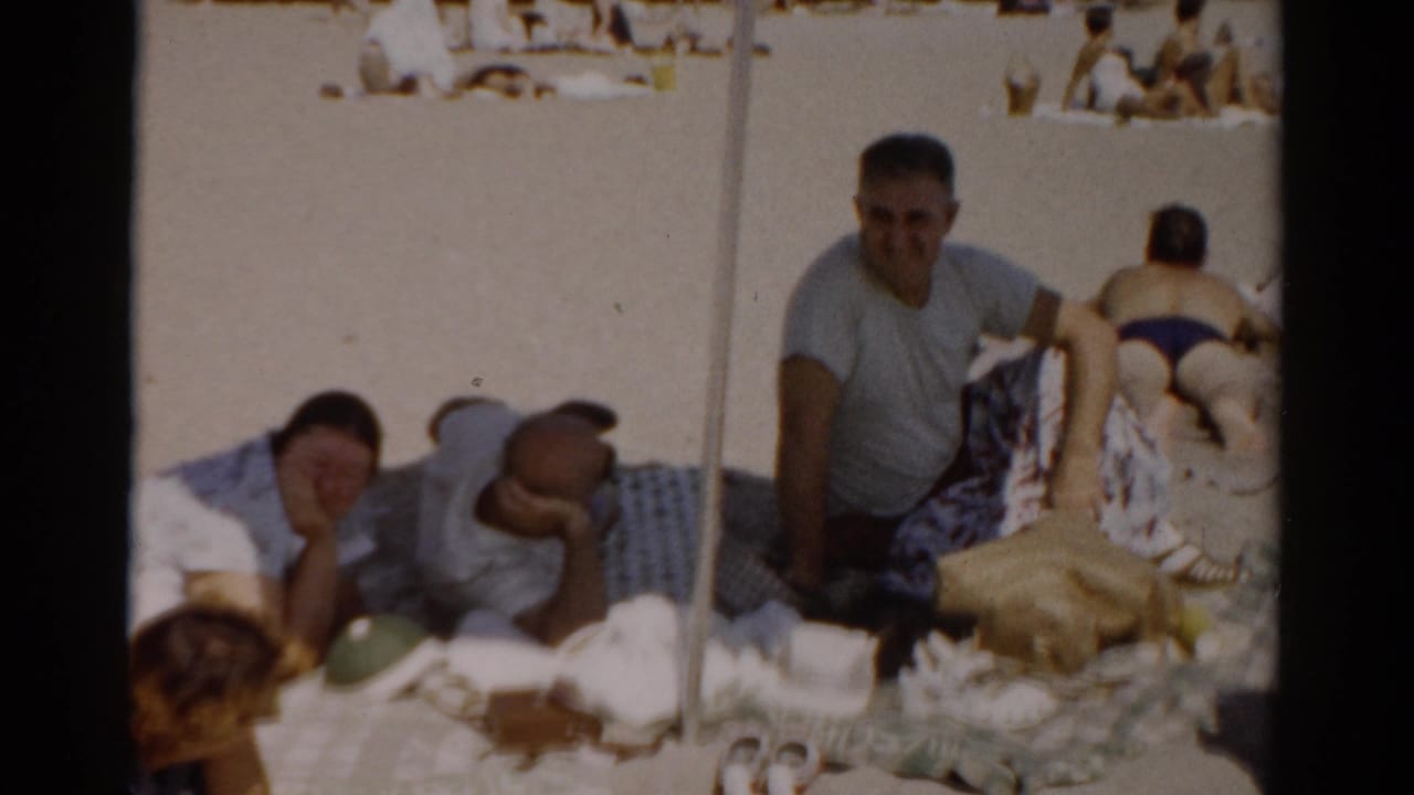 Man in grey t-shirt sitting upright and smiling on a beach blanket, woman with dark hair lying beside him, person in dark swimsuit with back to camera at right.