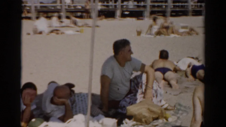 Man in grey t-shirt seated on a sunny sandy beach surrounded by beach gear, additional person reclining in foreground; 8mm home movie, Catskill, New York, circa 1957.