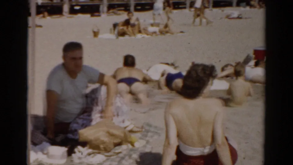 Man in grey t-shirt seated on sand with two women visible from behind, red container at frame edge; 8mm home movie, Catskill, New York, circa 1957.