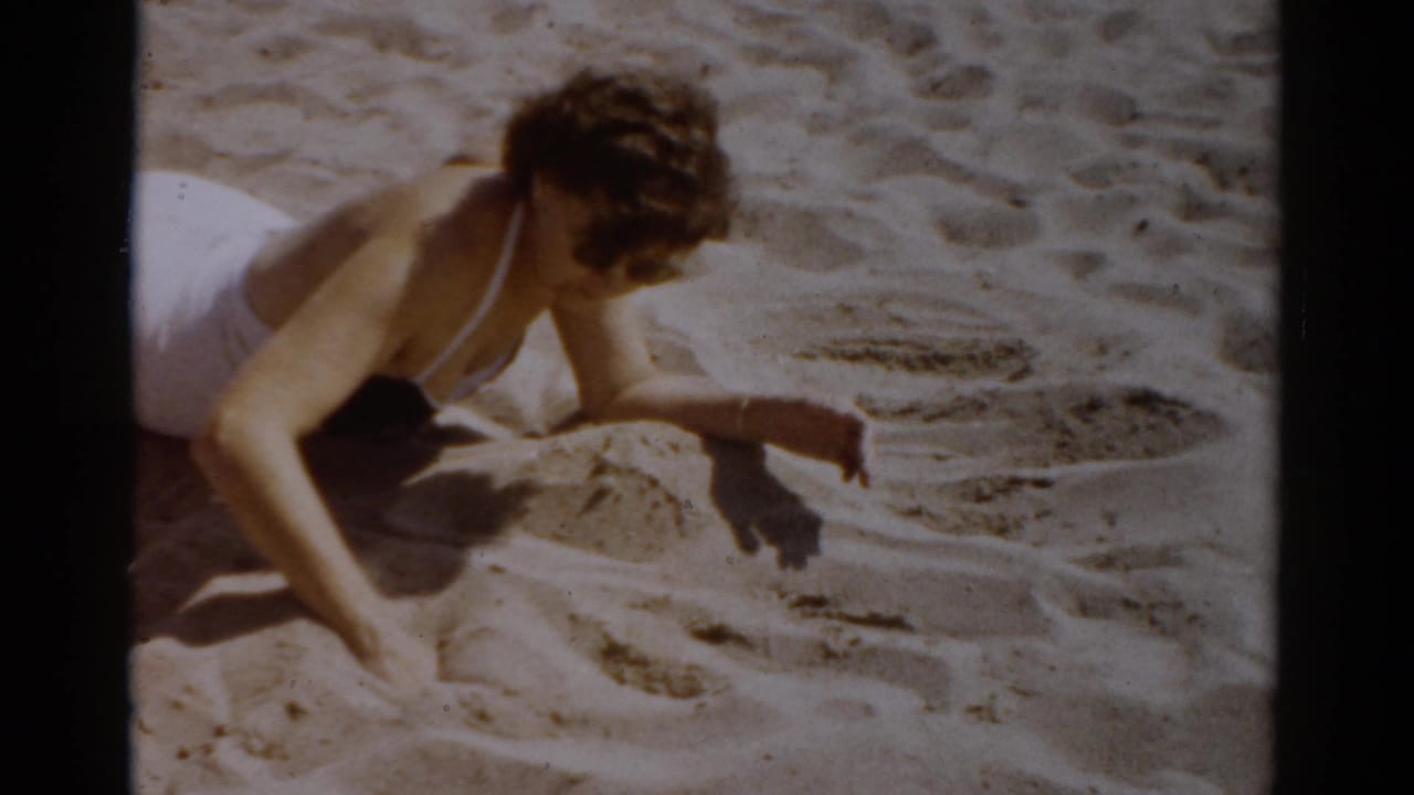 Woman in late-1950s white swimsuit kneeling on sandy beach, tracing a shape with her hand — 8mm home movie, c.1957, Catskill, New York.