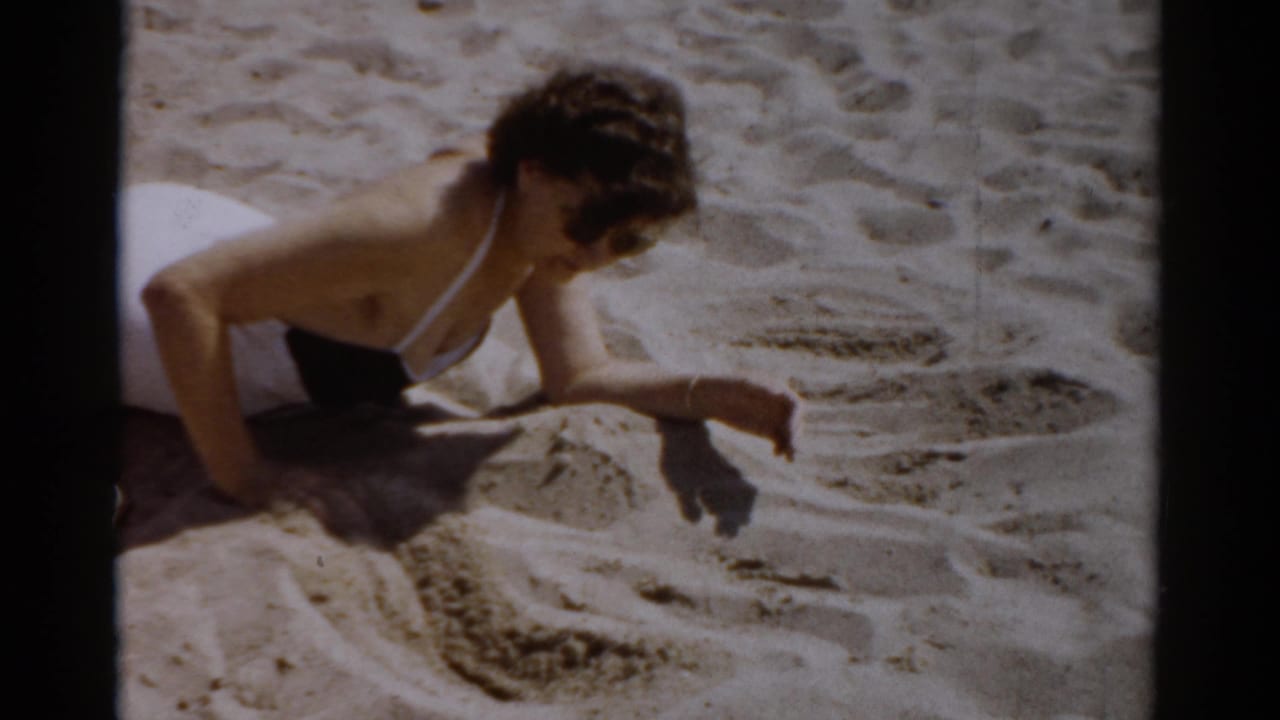 Woman in 1950s-style white swimsuit leaning over sandy beach, hand extended toward the sand — 8mm home movie, c.1957, Catskill, New York.