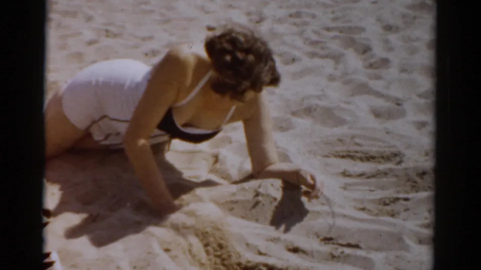 Woman in white 1950s-style bikini top crouching over sand, drawing with her finger — 8mm home movie, c.1957, Catskill, New York.
