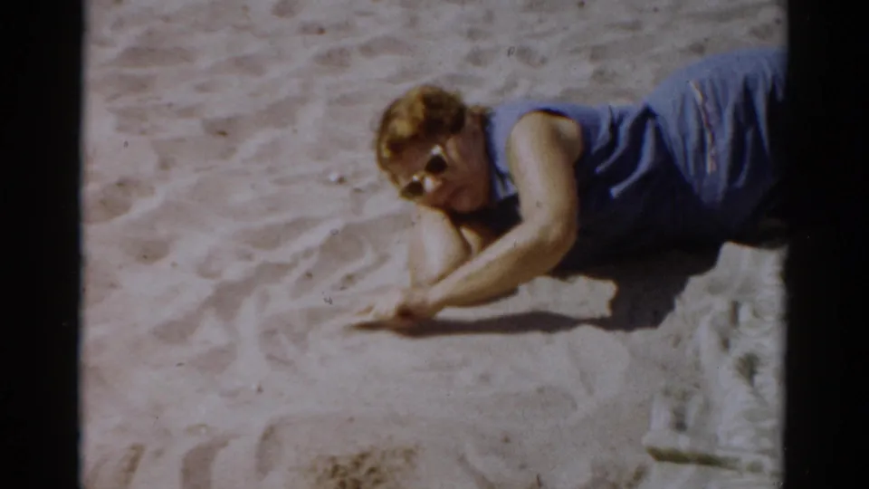 Woman in blue top and period dark-frame sunglasses lying on sandy beach, drawing shapes in sand — 8mm home movie, c.1957, Catskill, New York.