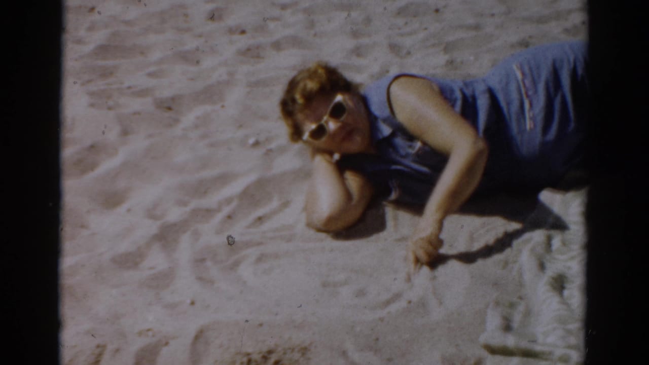 Young woman in blue sleeveless top and 1950s-style sunglasses reclines on beach sand tracing a pattern, in 8mm home movie footage from New York, 1957.