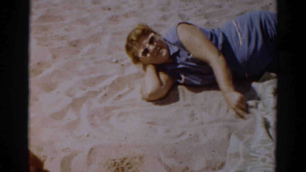 Same woman in blue sleeveless top reclining on sand, facing camera with sunglasses visible.