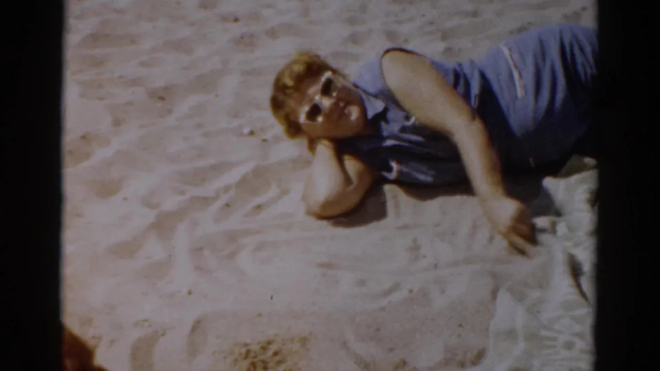 Close view of a woman in a blue sleeveless shirt reclining on beach sand and looking toward the camera, 1950s New York beach home movie, 1957.