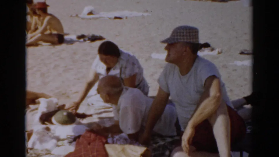 A man in a plaid bucket hat and a woman in a light dress sit together on a beach blanket with items spread around them; a small figure consistent with a child appears between them, New York beach, 1957.