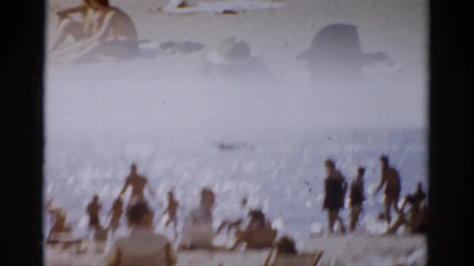Wide shot of a crowded summer beach with silhouetted bathers near the waterline, shot with warm hazy exposure on 8mm film, New York, circa 1957.