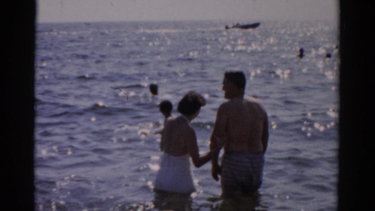 Man in striped trunks and woman in white one-piece swimsuit wade hand-in-hand in open water, 1957 8mm home movie; small boat on the horizon