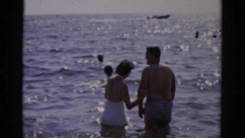 Man in striped trunks and woman in white one-piece swimsuit wade hand-in-hand in open water, 1957 8mm home movie; small boat on the horizon