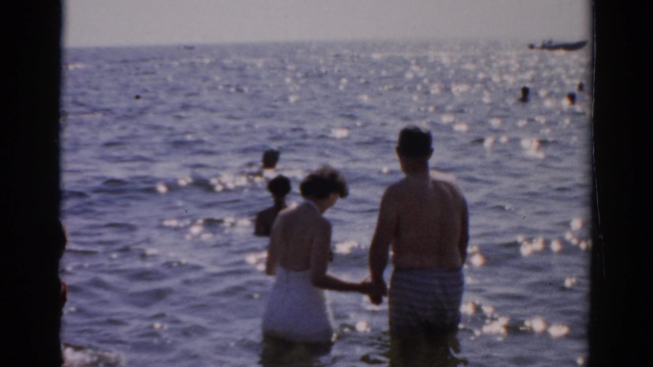 Man and woman in 1950s-era swimwear wade holding hands in open sunlit water; a small vessel appears upper right alongside scattered background bathers