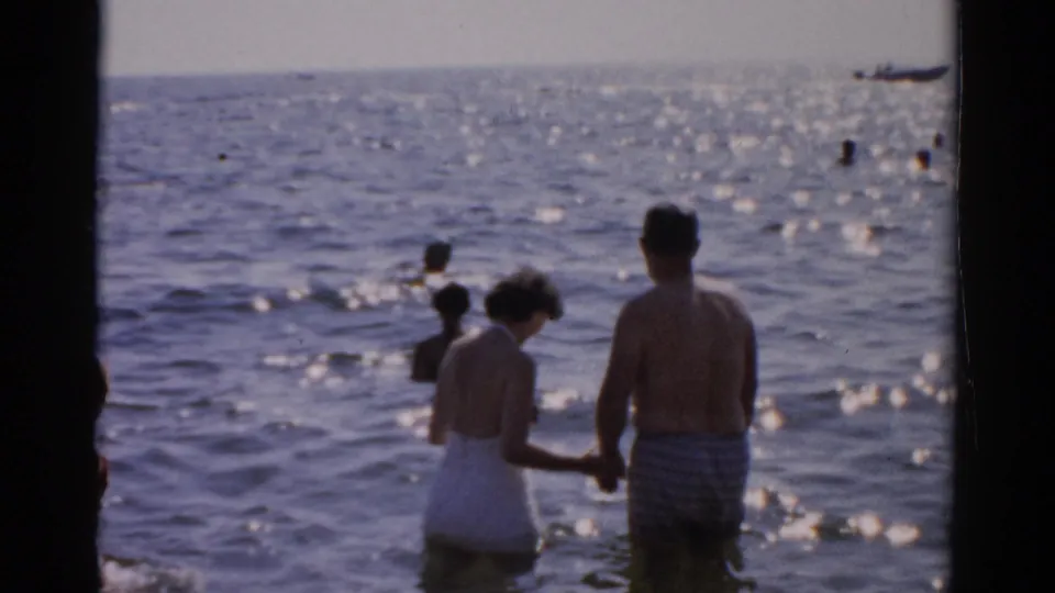 Man and woman in 1950s-era swimwear wade holding hands in open sunlit water; a small vessel appears upper right alongside scattered background bathers