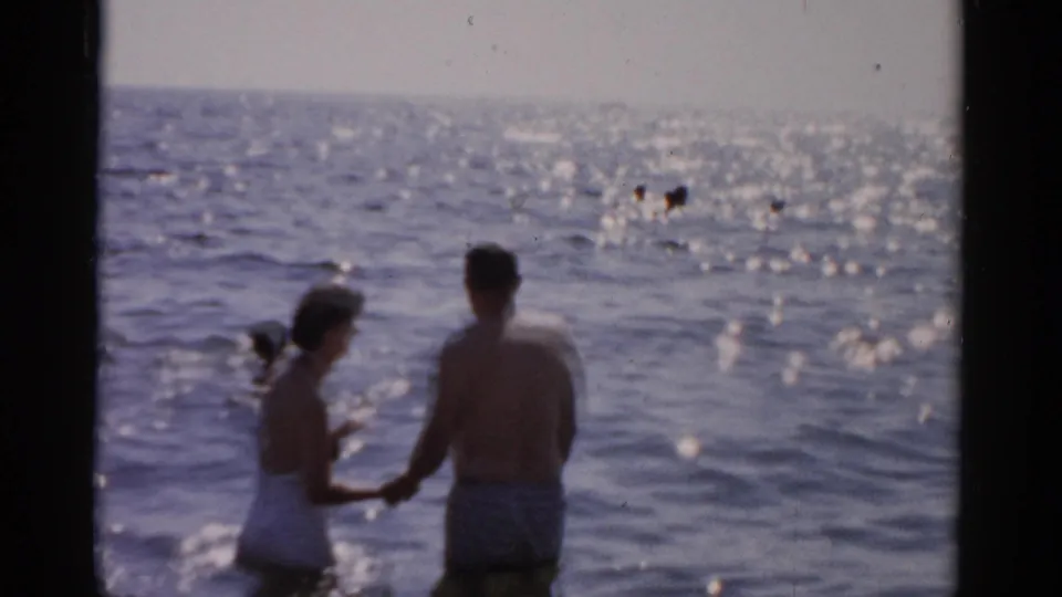 Man in striped trunks and woman in white swimsuit turn partly toward camera while wading together in broad sunlit ocean water