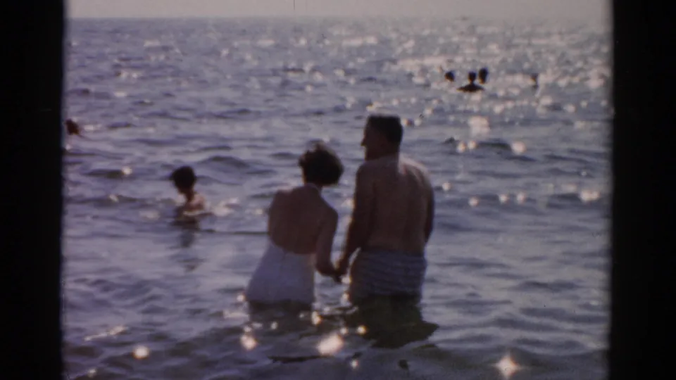 Man and woman in 1950s beachwear wade hand-in-hand while a swimmer appears in the middle distance and other bathers dot the sunlit water behind them