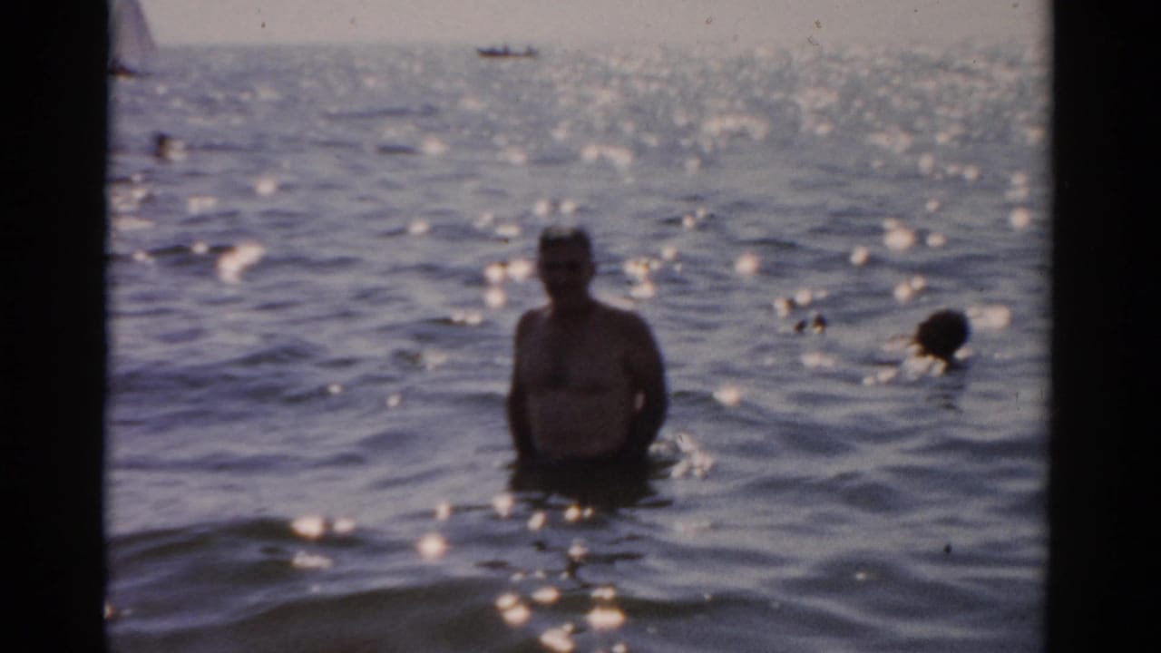 Adult man standing waist-deep in sunlit ocean, facing camera with arms partially raised; 1950s 8mm home-movie footage, location metadata records Catskill, New York.