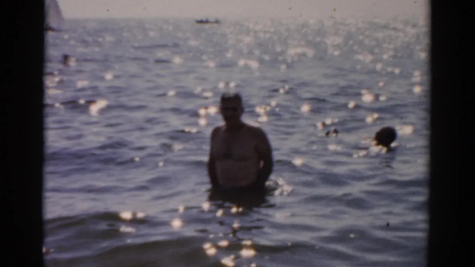 Adult man standing waist-deep in sunlit ocean, facing camera with arms partially raised; 1950s 8mm home-movie footage, location metadata records Catskill, New York.