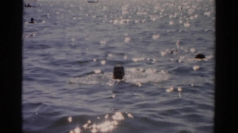 Scattered swimmer figures in choppy ocean water with strong sun-sparkle reflections; no central foreground subject; 1950s 8mm home-movie footage.