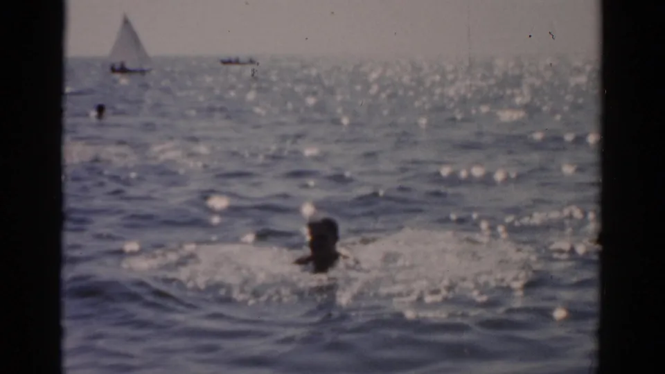 Swimmer in lower foreground with a white-sail sailboat on the horizon and scattered bathers beyond; 1950s 8mm home-movie footage at a coastal or large-lake setting.