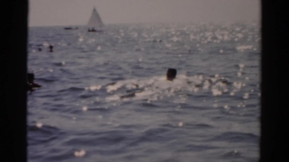 Swimmer in right foreground on open ocean with a white-sail sailboat prominently on the horizon; 1950s 8mm home-movie coastal footage.