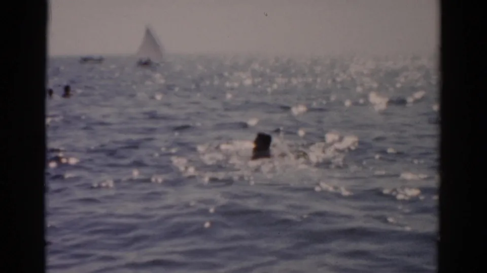 8mm film frame of multiple swimmers in open water with a distant sailboat; New York coast, circa 1957.