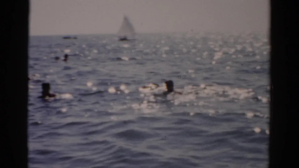 8mm film frame of swimmers in sun-lit open water with a sailboat in the distance; New York coast, circa 1957.