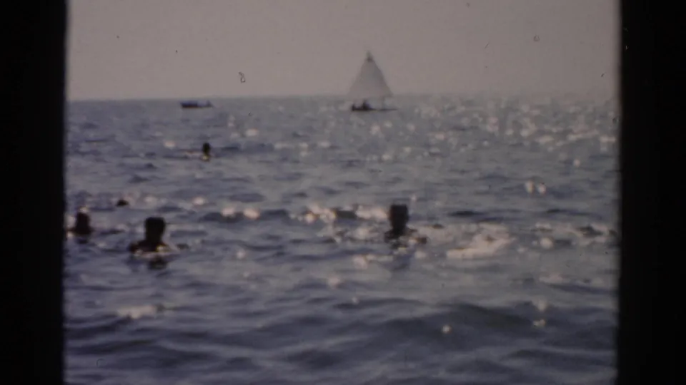 8mm film frame of multiple swimmers in ocean water with a white sailboat under sail in the mid-distance; New York coast, circa 1957.