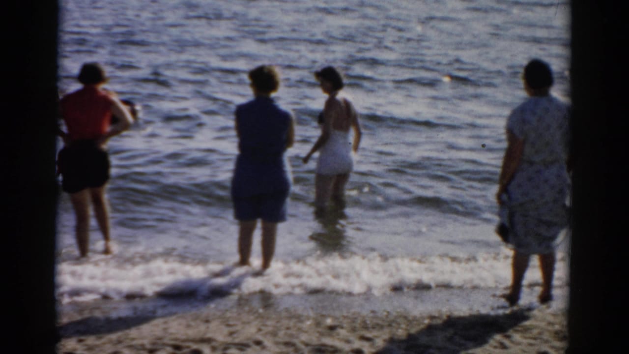 Several women in mid-century swimwear and casual dresses stand at the water's edge, viewed from behind, as small waves break on a sandy shore.