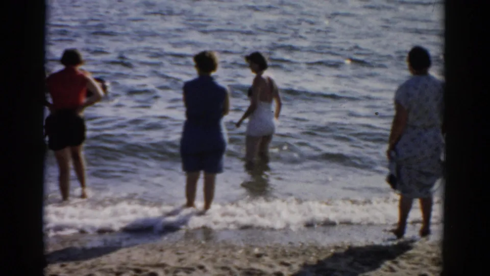 Women in late-1950s swimsuits and summer dresses wade at a sandy waterfront, figures facing the water, circa 1957, New York.