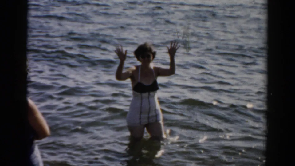 A woman in a mid-century dark swimsuit waves with both hands raised at the camera while standing waist-deep in water, circa 1957.