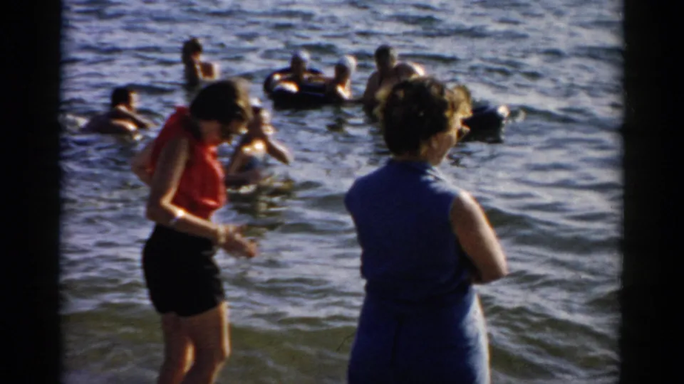 Foreground women in mid-century summer attire near the water; background group swims with an inflatable ring, circa 1957, New York waterfront.
