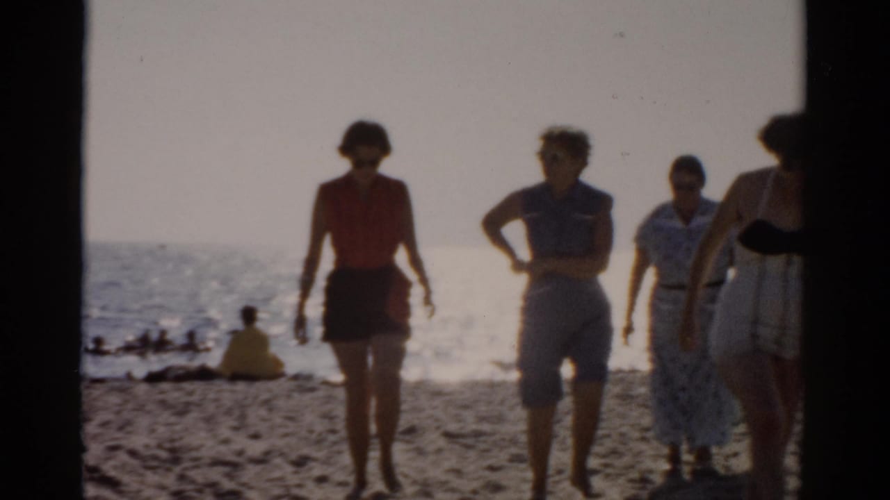 Four women in mid-1950s casual beachwear, including a red sleeveless top with high-waisted shorts and a patterned dress, walk toward the camera on a bright sandy beach, circa 1957.
