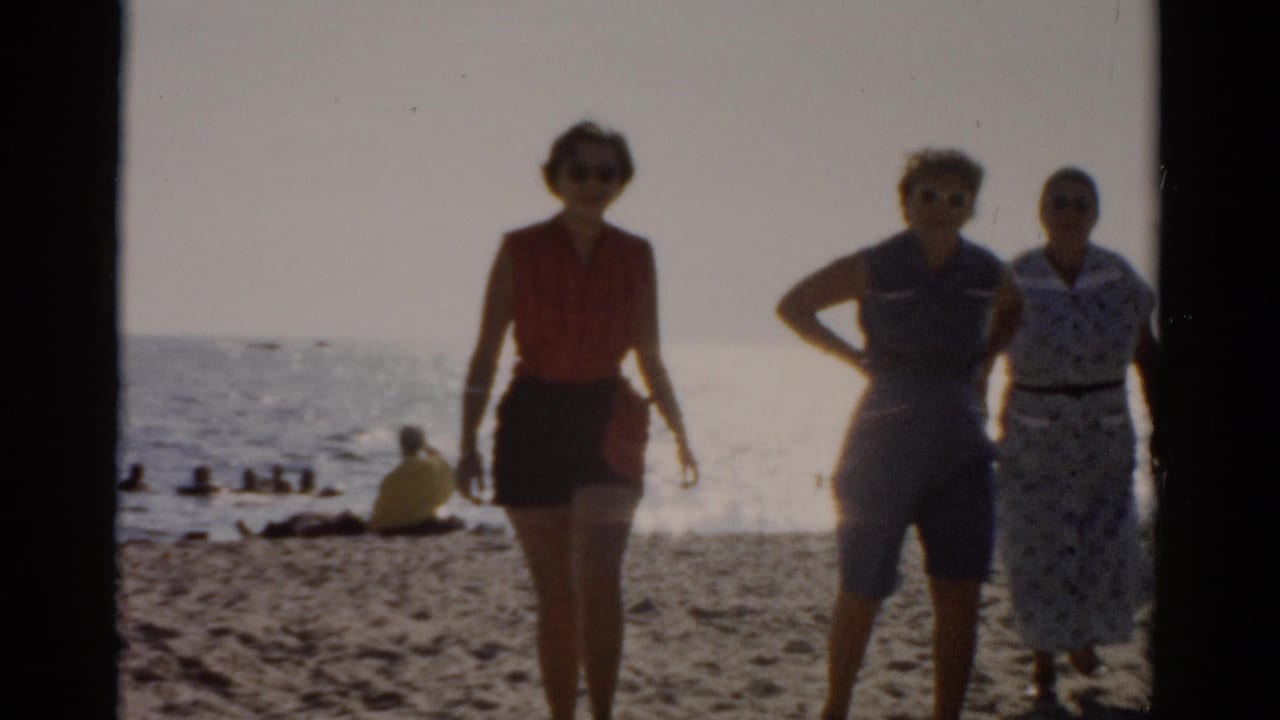 Three women in 1950s summer attire face the camera on a sandy beach; ocean and distant seated bathers visible behind them.