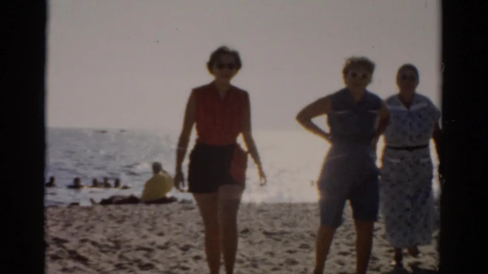 Three women in 1950s casual beach dress, including a red top with dark shorts, a light blue outfit, and a patterned dress, face the camera on a sandy beach with ocean behind them, circa 1957.