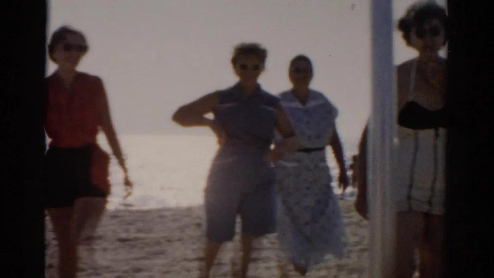 Three or four women in 1950s casual attire walk and gesture playfully toward the camera on a bright sunny beach; film sprocket border visible at the left edge of the frame, circa 1957.