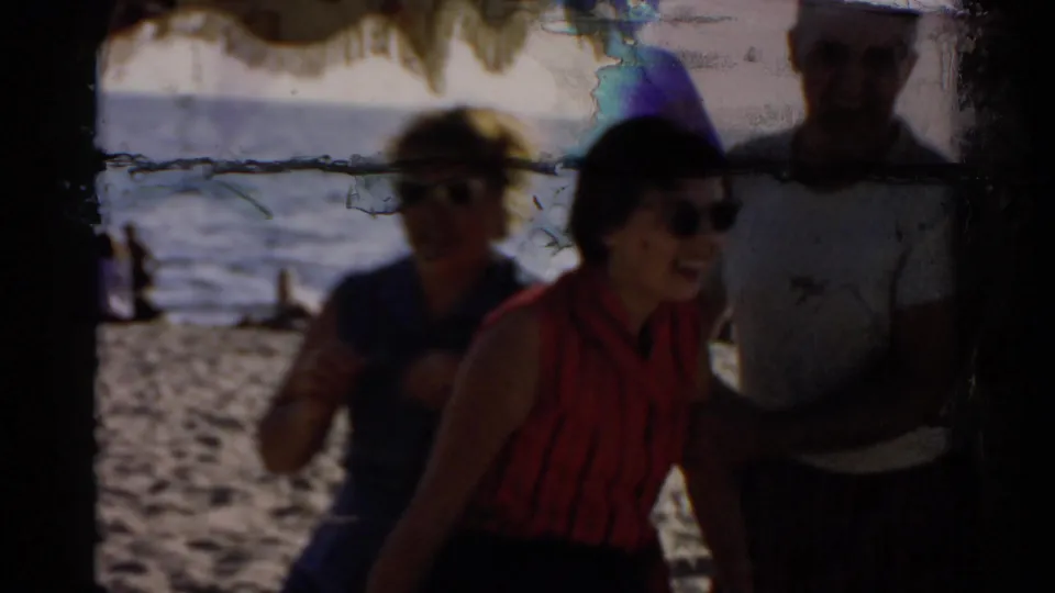 Tight two-shot of two women wearing sunglasses, one in a red top, laughing on a sunny beach; chromatic emulsion damage streaks visible at top of frame, circa 1957.