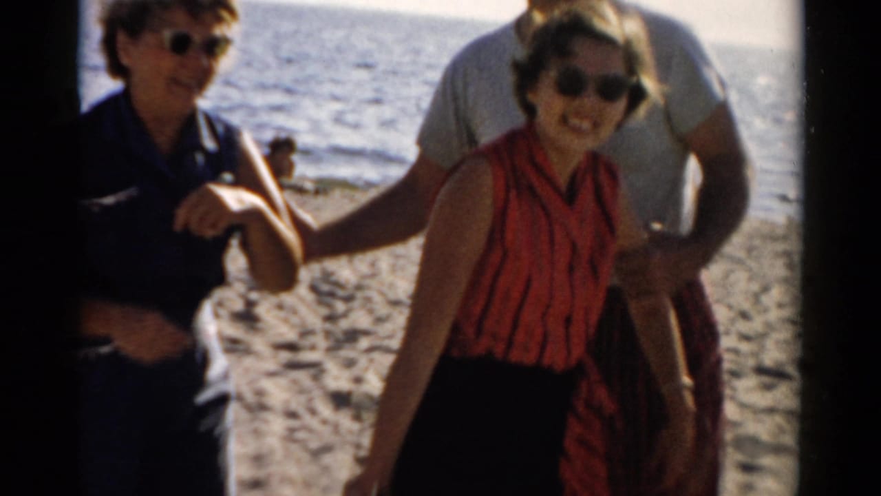 Two women and a man on a sandy beach; woman in red blouse and sunglasses at center smiling, second woman in blue top and cat-eye sunglasses at left, man in white shirt behind center.