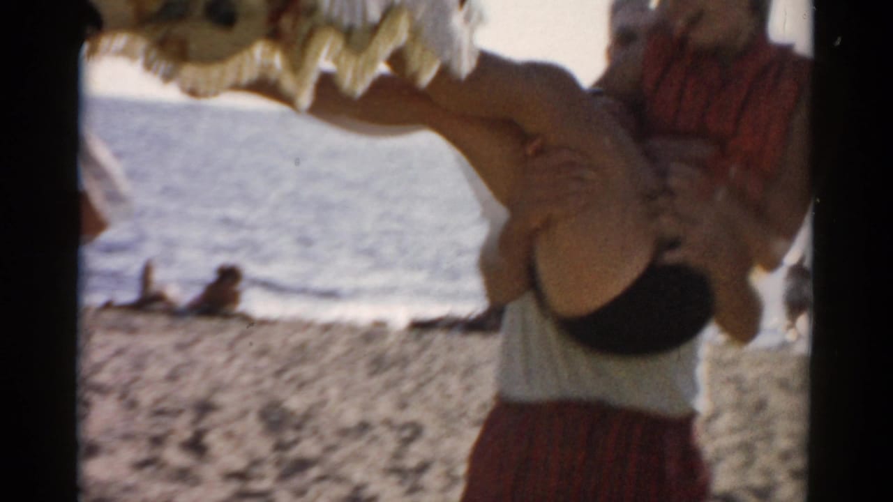 Man in red plaid shorts begins lifting a woman overhead on a sandy beach, ocean visible behind; thatched or palm-frond structure partially visible at top of frame.