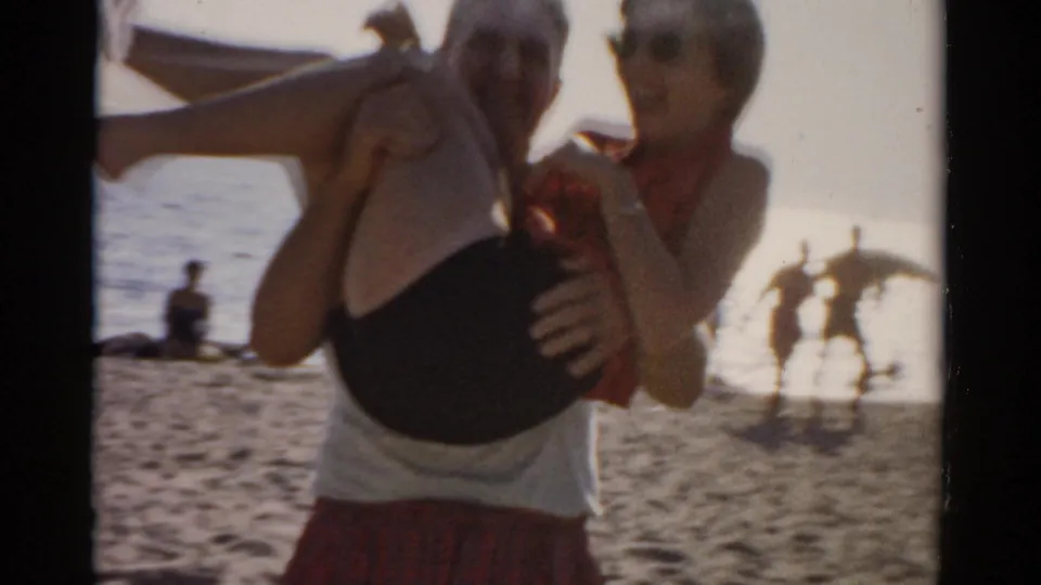 Man in white undershirt and red plaid shorts cradles a woman in a black swimsuit on a sunny beach, late 1950s 8mm; what appear to be large birds with spread wings are visible in the background midground.