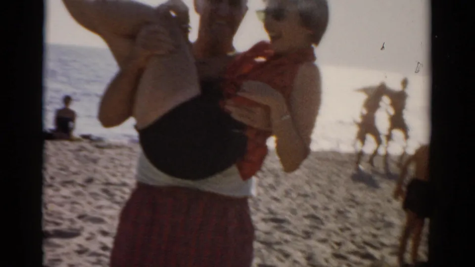 Man holds a woman on a sun-drenched beach in the late 1950s, both in mid-century leisurewear; background shows standing figures, bird-like shapes, and water on 8mm color film.