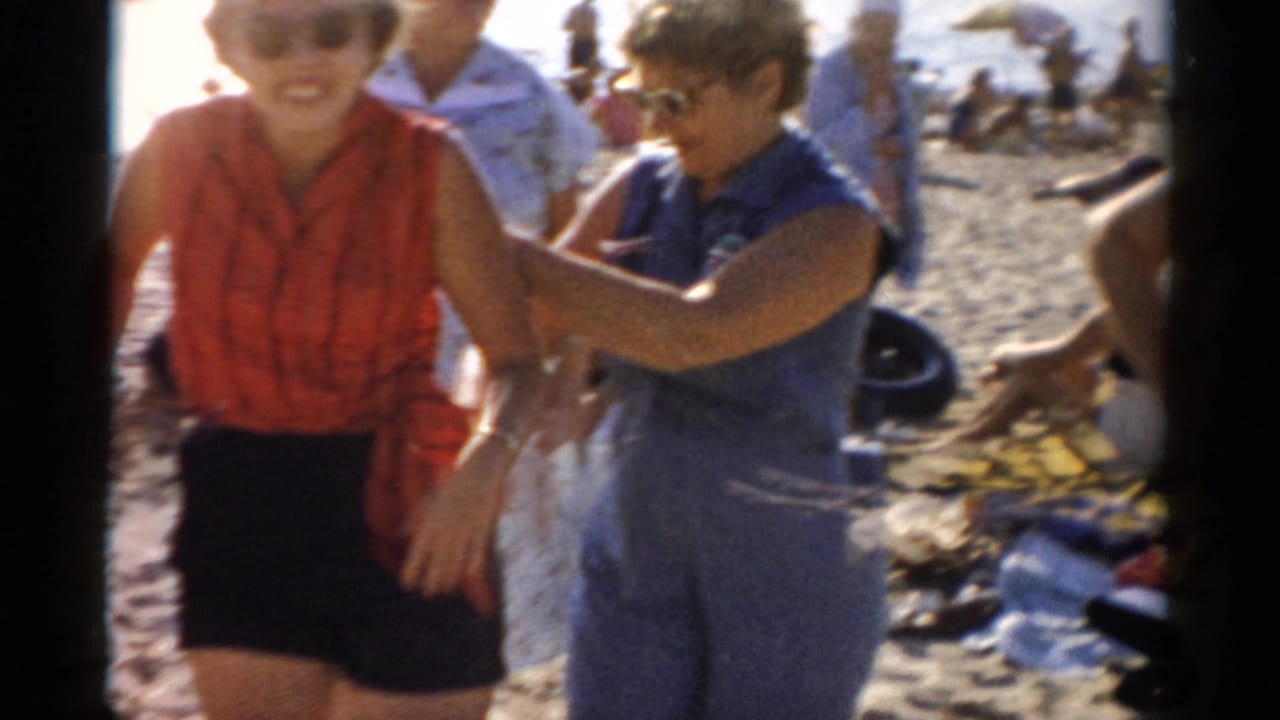 Two women, one in a red sleeveless top and black shorts, one in a blue dress, laugh and jostle playfully on a busy beach in what metadata dates to 1957 in New York state.