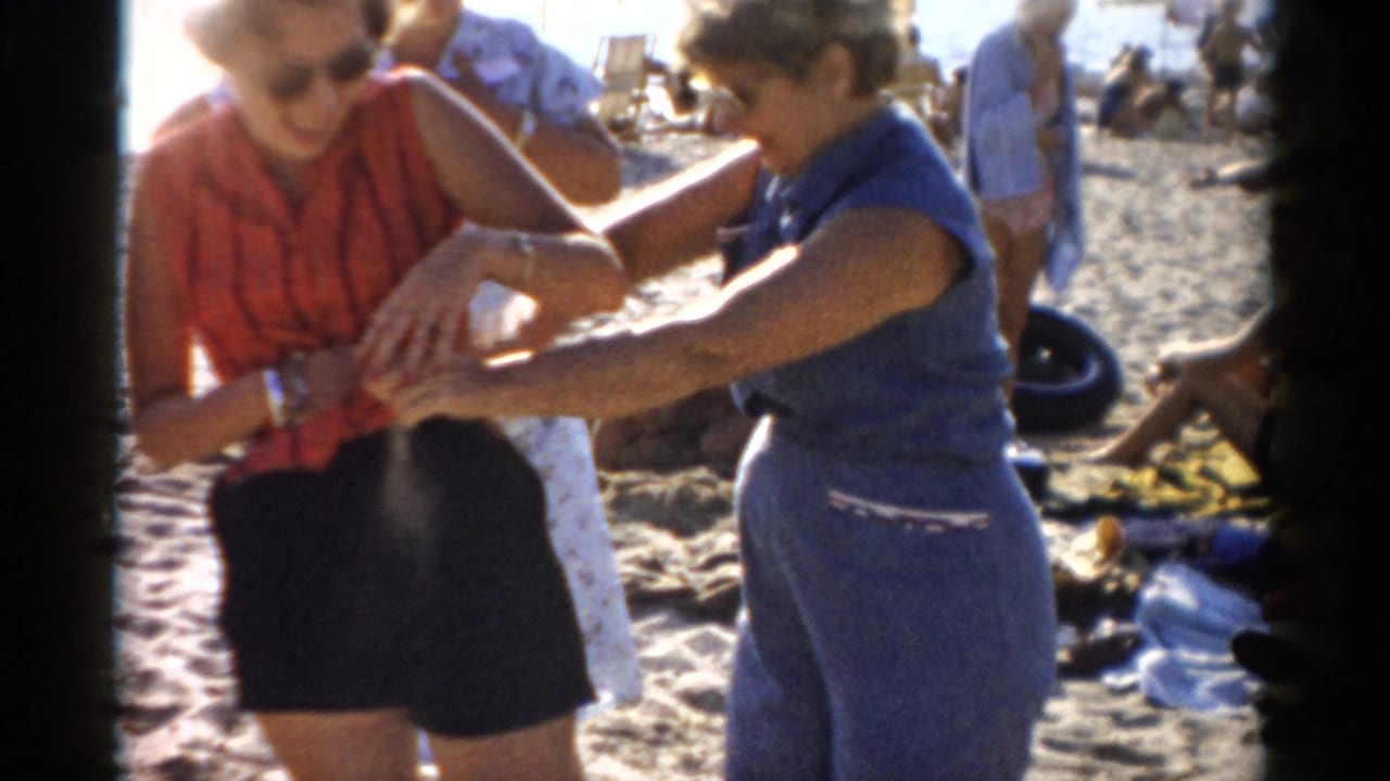 Two women in colorful summer clothing grabbing or clasping each other playfully on a sandy beach.