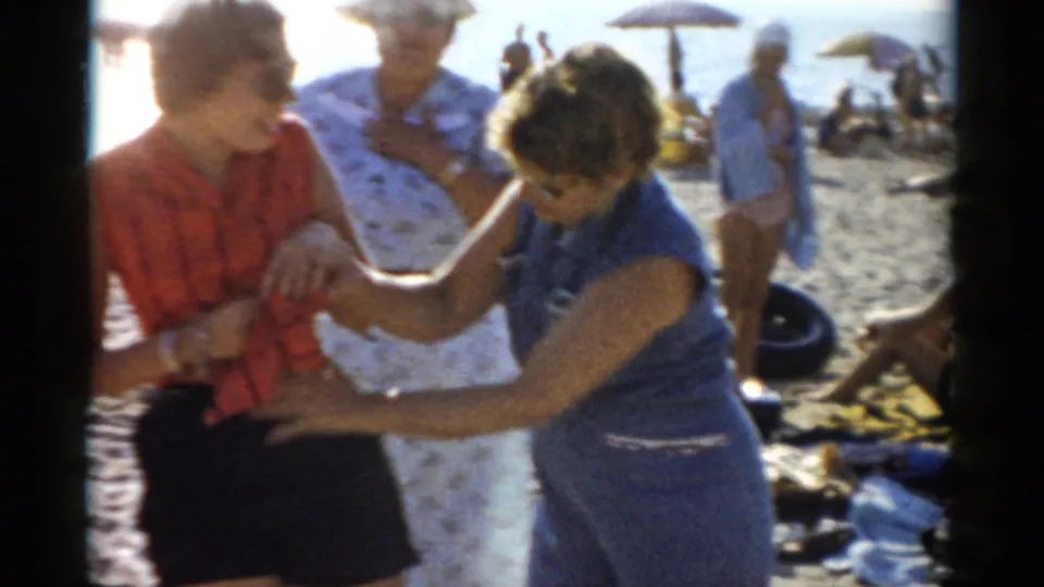 The two women continue their interaction on a crowded 1950s beach; a striped beach umbrella, a solid umbrella, and a man in a light blue shirt are visible in the background.