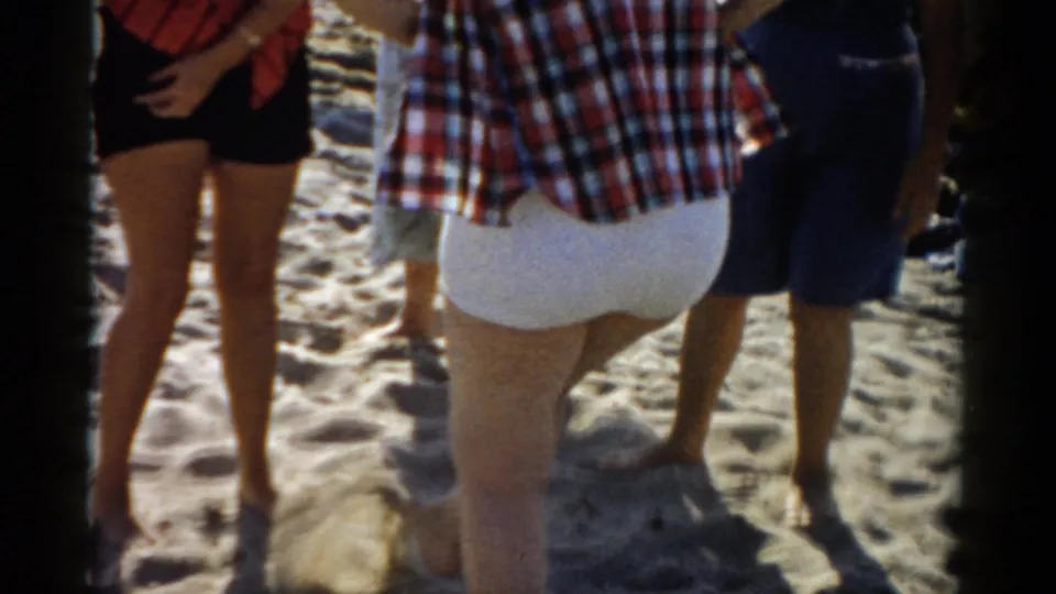 Three figures shot from waist level on a sandy beach; visible clothing includes black shorts with a red top, white shorts, and a red plaid shirt consistent with 1950s casual summer wear.